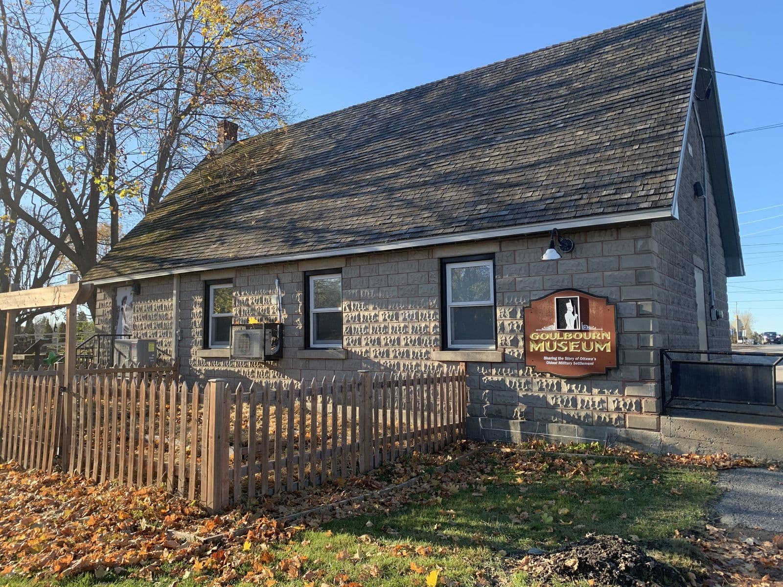 Le bâtiment en blocs de pierre du musée Goulbourn, à Stittsville, derrière une clôture de piquets en bois, avec des feuilles d’automne sur la pelouse sous un ciel bleu