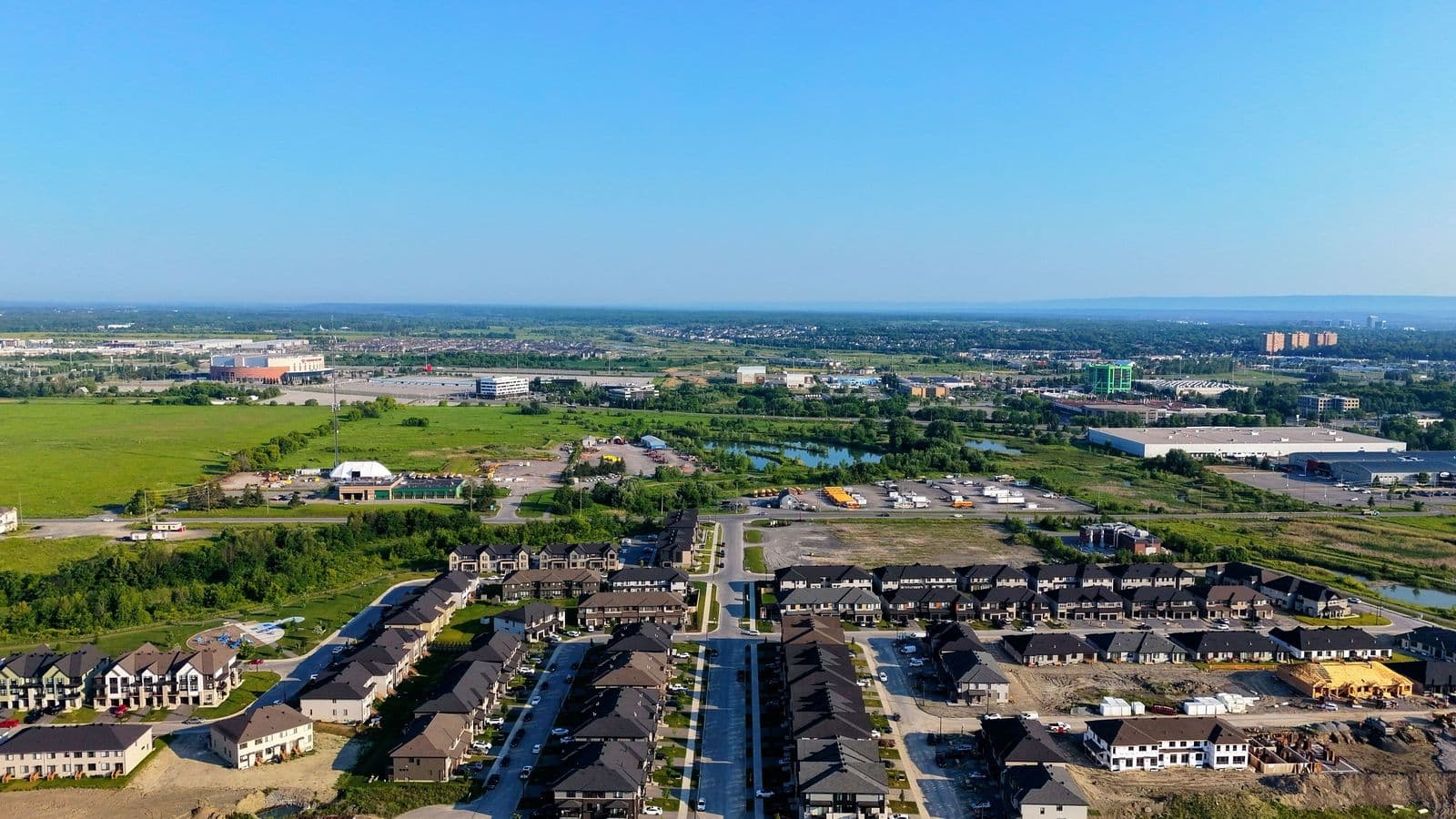 Vue aérienne de nouvelles maisons en rangée dans des rues fraîchement asphaltées de l’ouest de Kanata, avec des champs, un étang et l’aréna du Centre Canadian Tire au loin