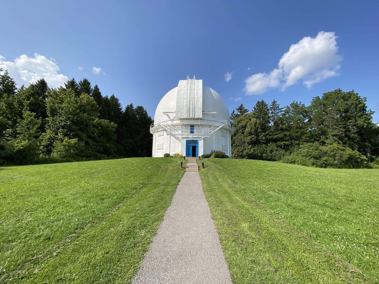 The white telescope dome of the David Dunlap Observatory in Richmond Hill at the end of a gravel path across a mown summer lawn, framed by evergreens under a blue sky