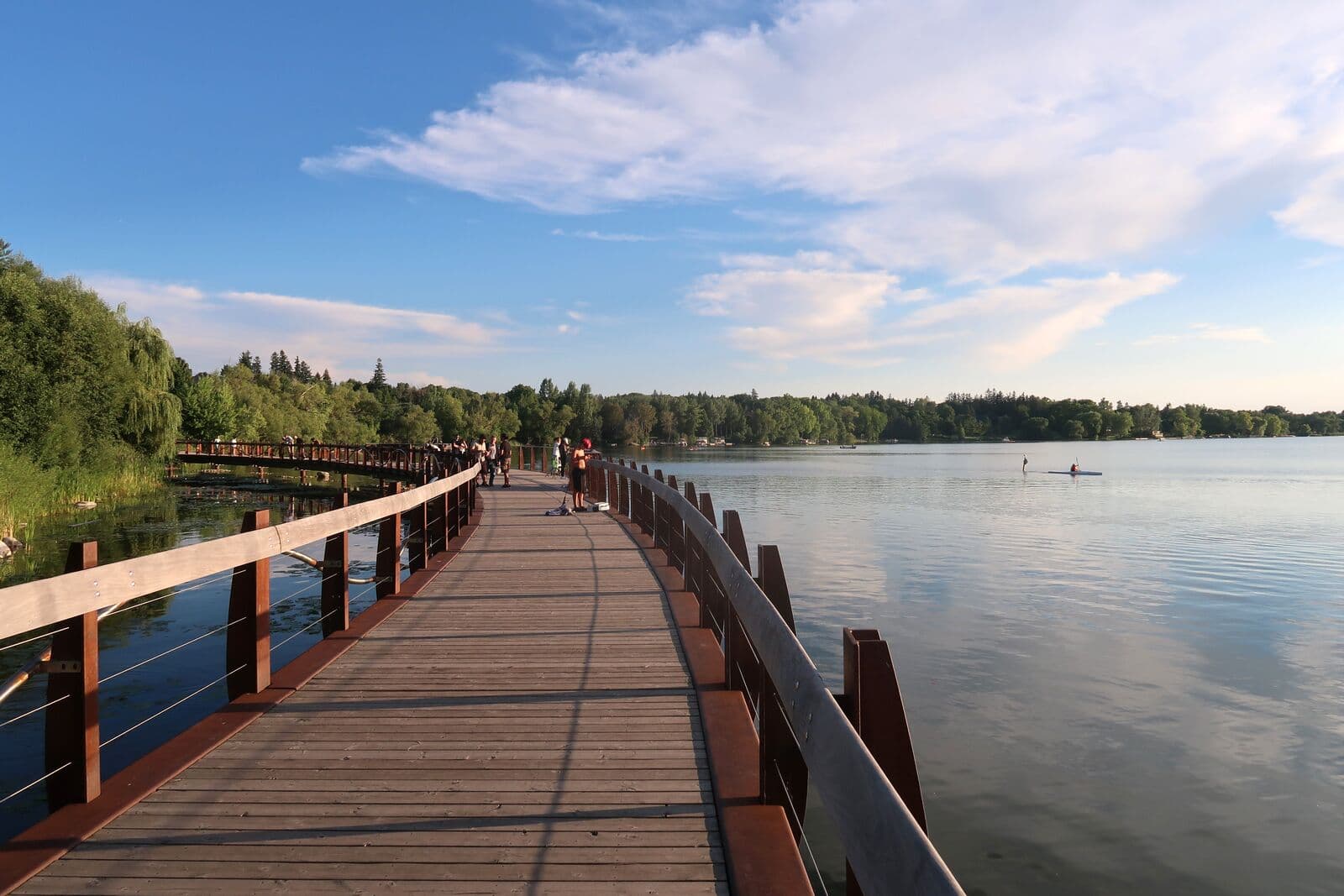 A curving wooden boardwalk over Lake Wilcox on a summer evening, with walkers in the distance and a paddleboarder on the calm water