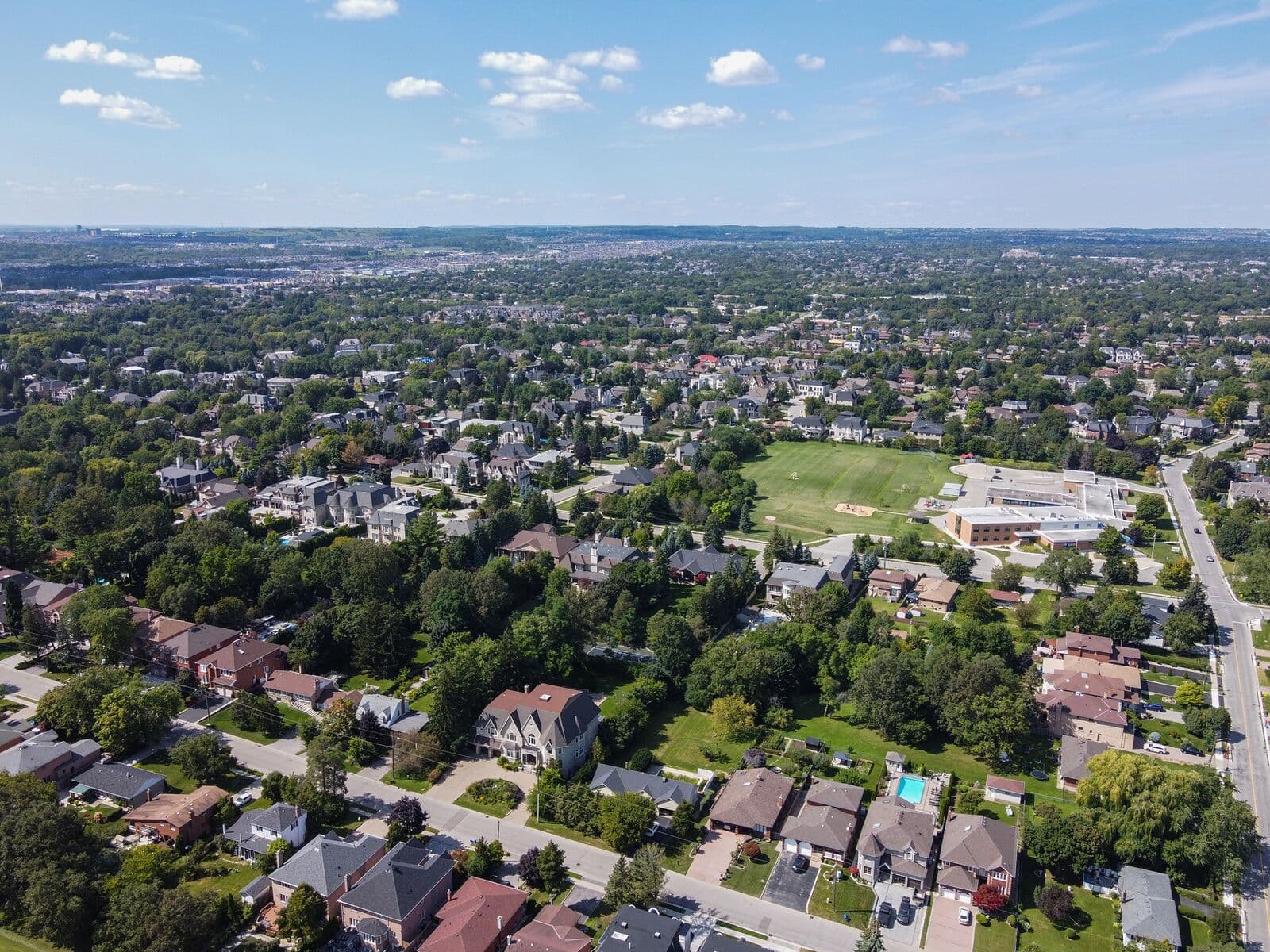 Aerial summer view over leafy streets of detached houses in the Richvale neighbourhood of Richmond Hill, with a school and its playing field and suburbs stretching to the horizon