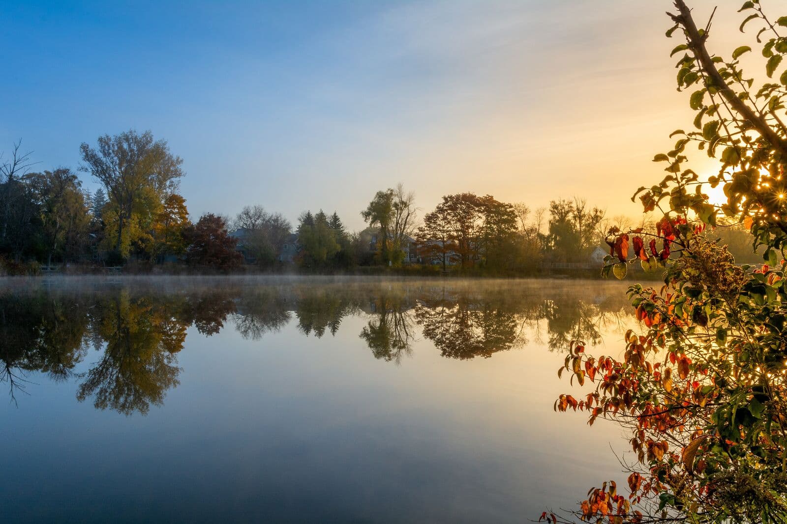 Sunrise over a still, misty Toogood Pond in Unionville in autumn, with fall trees mirrored in the water and red leaves in the foreground