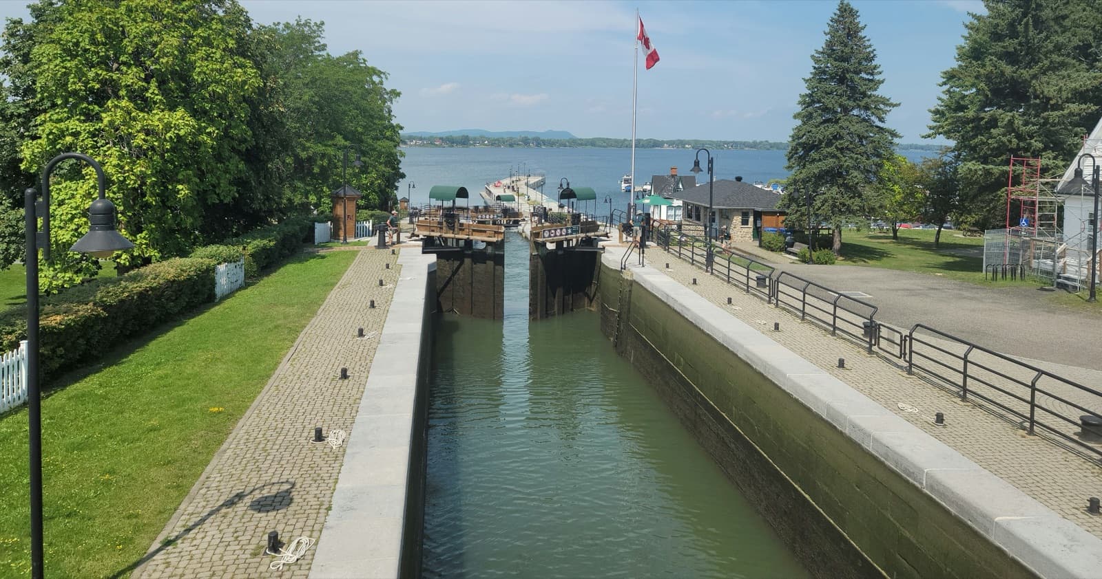 A stone lock of the Chambly Canal in Chambly with its wooden gates opening, cobbled lockside paths and a Canadian flag, the Chambly basin beyond
