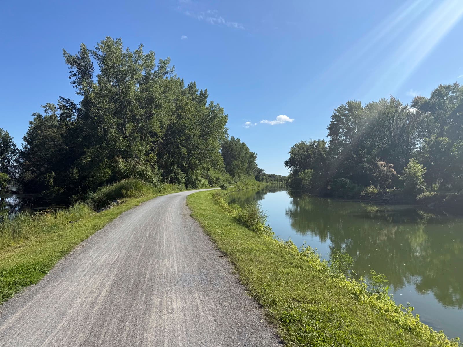 The gravel towpath of the Chambly Canal curving between leafy trees beside still water that mirrors them, on a sunny summer afternoon