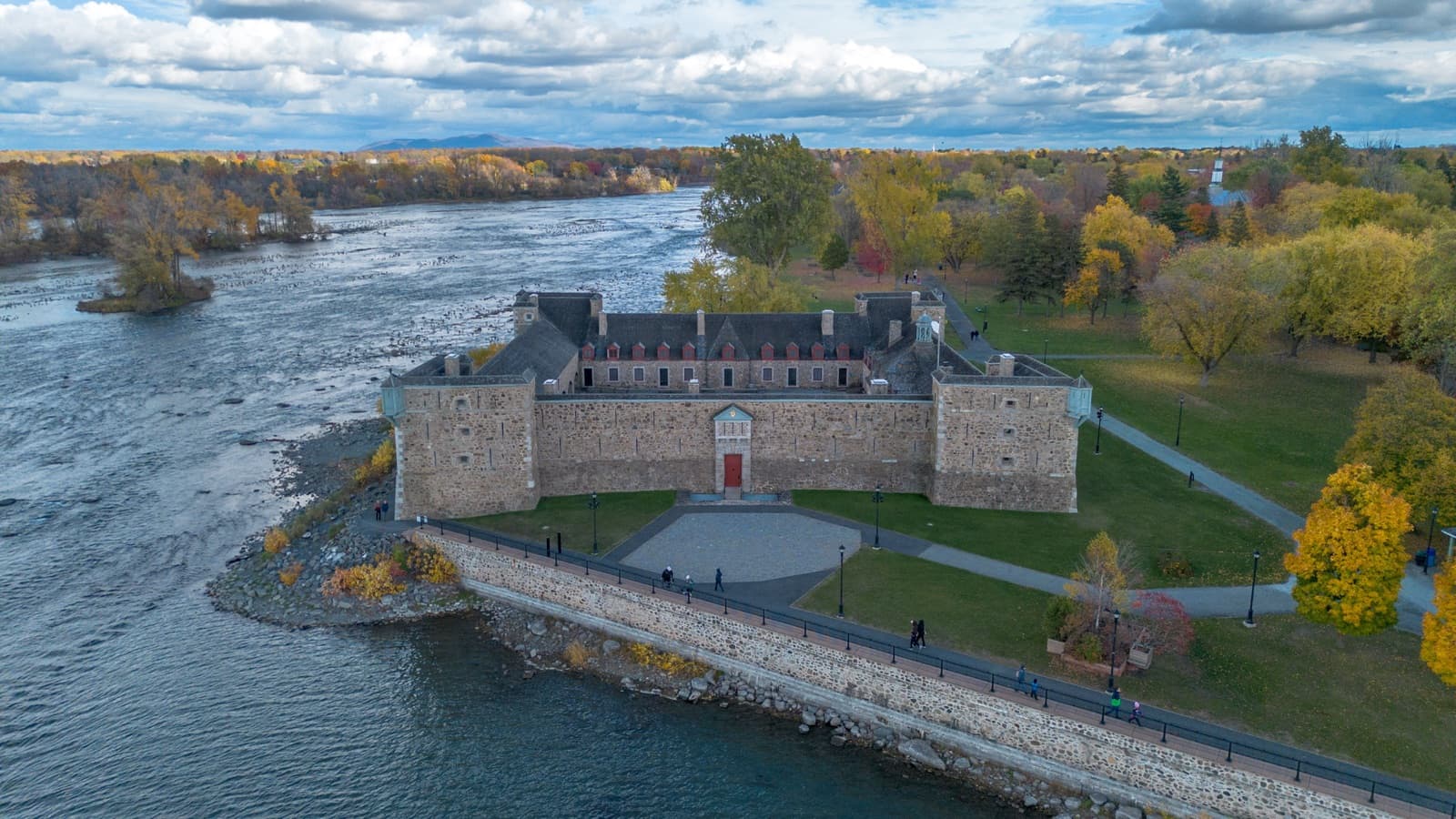 Fort Chambly’s stone walls on the Richelieu at the foot of the rapids, with autumn trees, lawns and a riverside promenade, seen from above