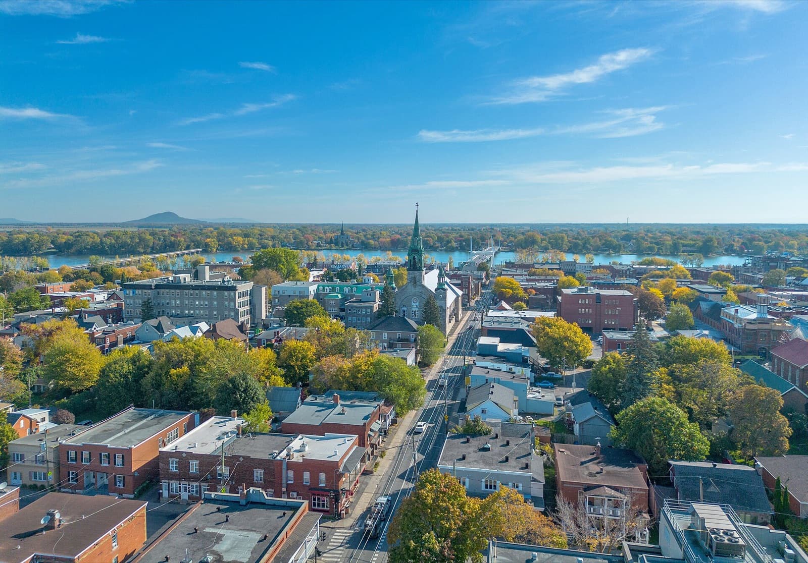 Brick buildings and autumn trees of downtown Saint-Jean-sur-Richelieu leading to the cathedral’s green spire, with the Richelieu River and Iberville on the far bank