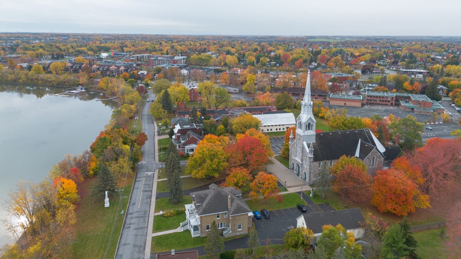Old Chambly in peak fall colour from above, with a stone church and its spire, red and orange maples and a street along the Chambly basin