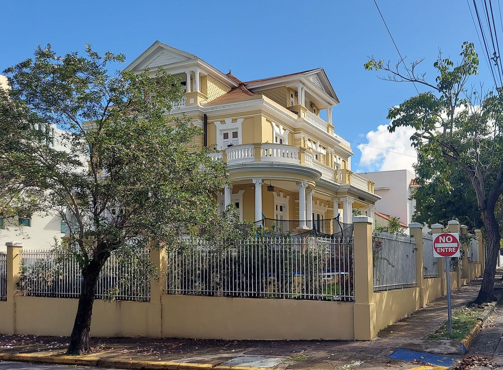 Una casa amarilla de tres pisos con columnas blancas, balcones redondos y techo de tejas rojas detrás de una verja de hierro en una esquina de Miramar
