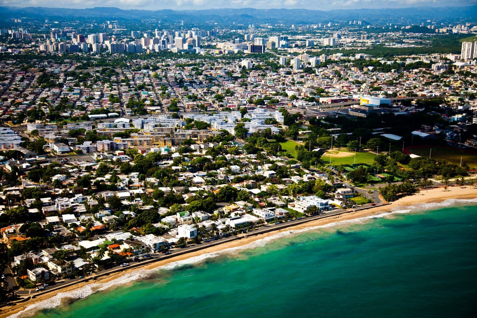 Vista aérea de Ocean Park: casas bajas y palmas junto a una playa de arena y agua verde, con canchas a la derecha y el perfil de San Juan al fondo