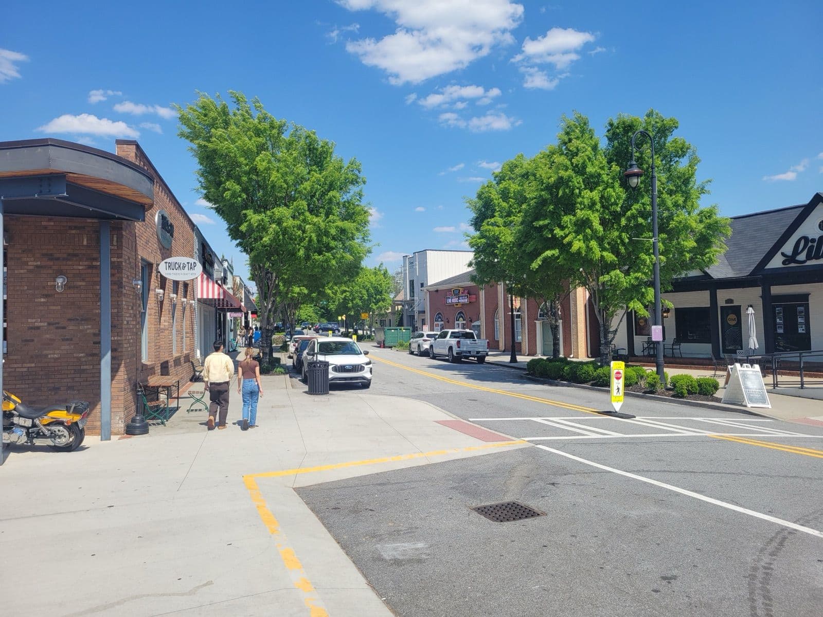 Tree-lined Main Street in downtown Duluth with brick storefronts, sidewalk seating and a live music marquee on a sunny spring day