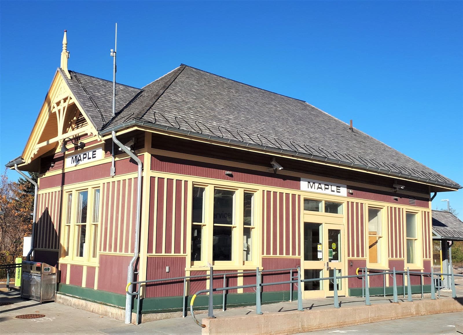 The maroon-and-cream wooden Maple GO station building from 1903, with its carved gable, MAPLE name boards and a slate-grey roof under a clear blue sky
