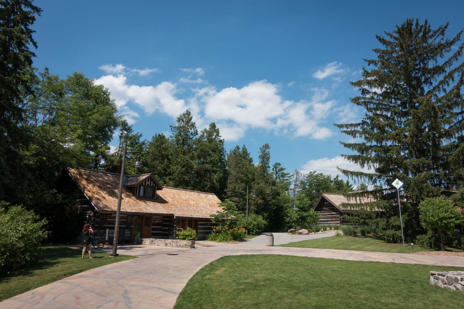 Log buildings with cedar-shingle roofs among tall pines and lawns on the McMichael Canadian Art Collection grounds in Kleinburg, under a blue summer sky