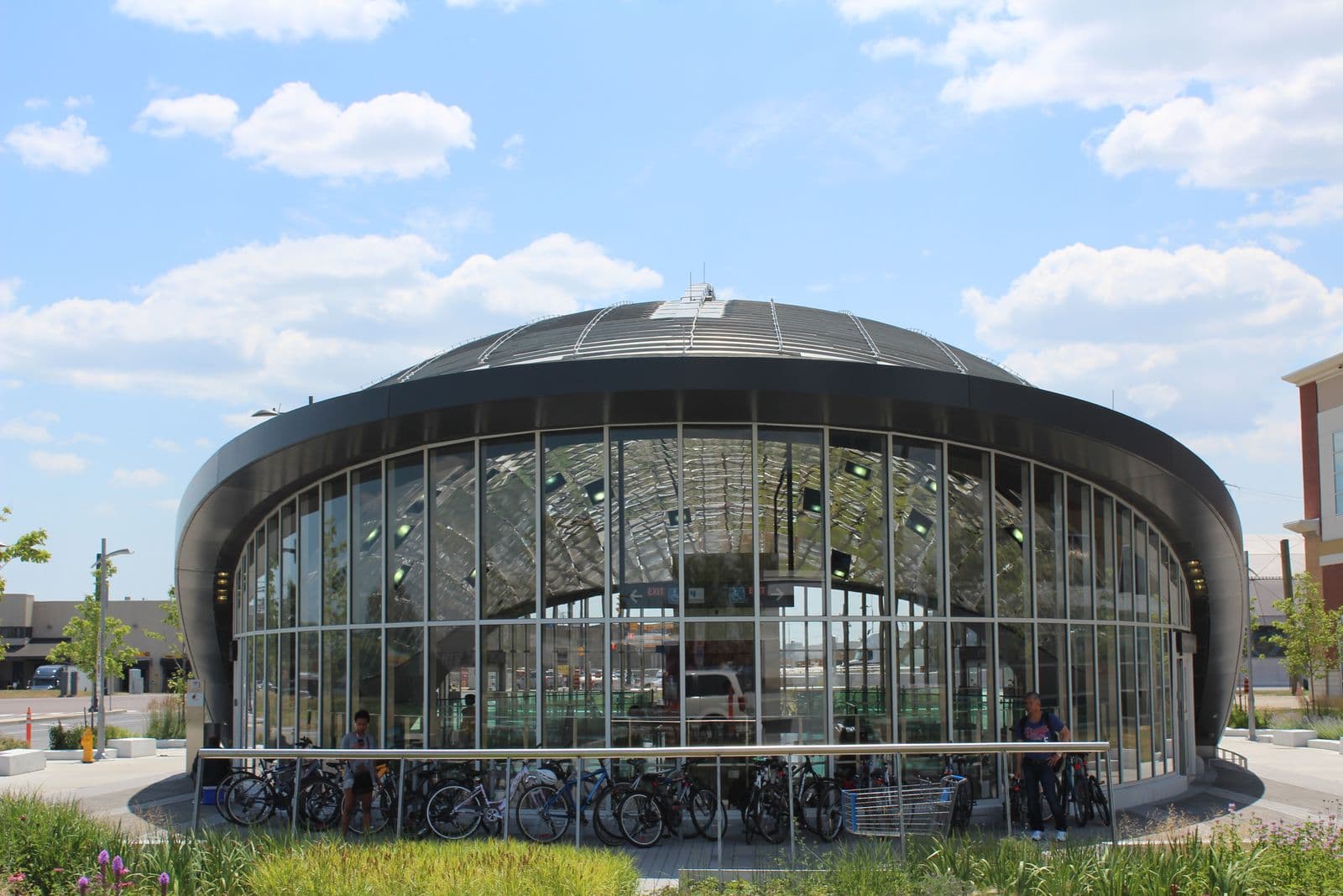 The domed glass entrance of Vaughan Metropolitan Centre subway station, with bicycles parked along its railing and summer clouds overhead
