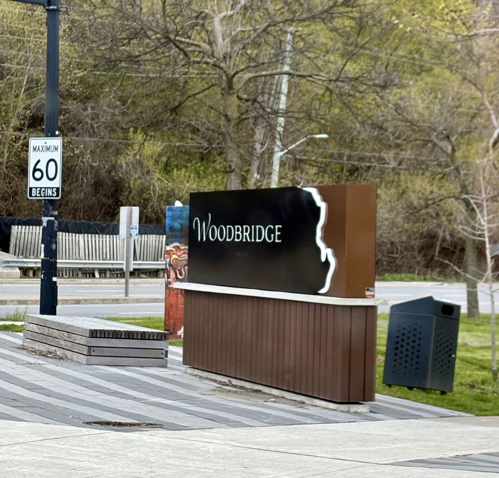 The dark Woodbridge gateway sign beside a bench and a painted traffic-signal box at Woodbridge Avenue and Islington Avenue, with spring trees on the Humber valley slope behind