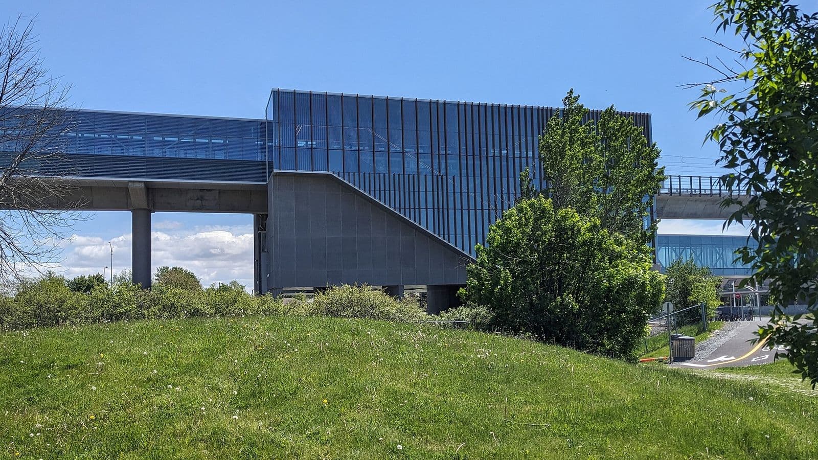 La station Kirkland du REM, en verre et en béton sur sa voie surélevée, vue au-delà d’une butte gazonnée du parc des Bénévoles sous un ciel bleu