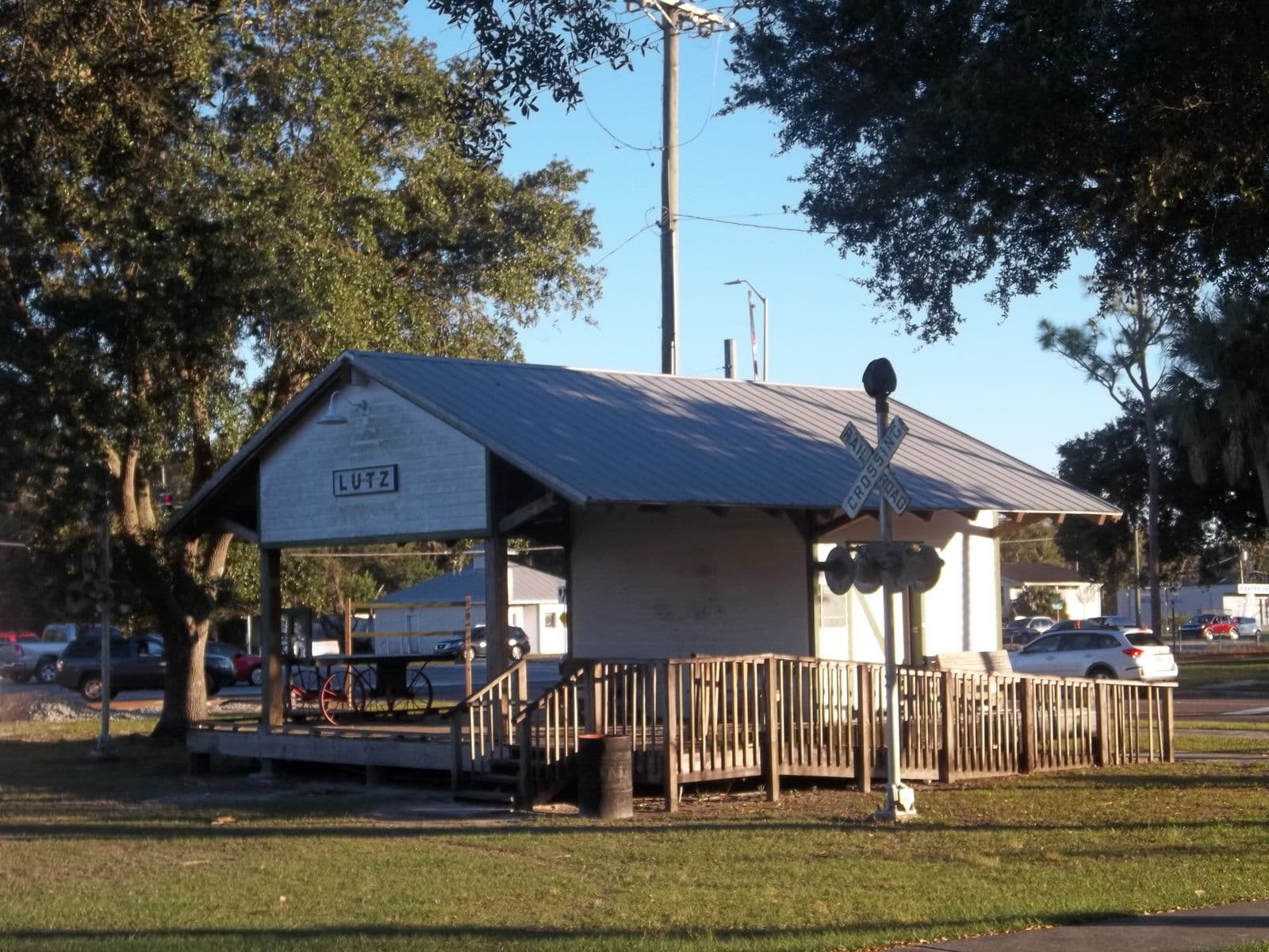 The white wooden Lutz depot replica with its Lutz sign, a railroad crossing signal and big oak trees in downtown Lutz under a clear blue sky