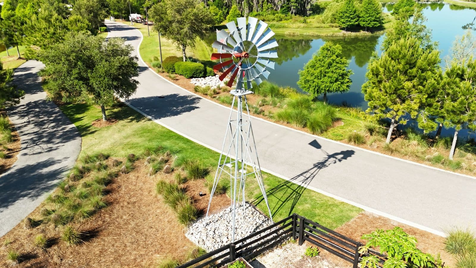 A farm-style windmill beside a curving paved path and a calm, tree-lined lake at Cunningham Park in Odessa, seen from above on a sunny day