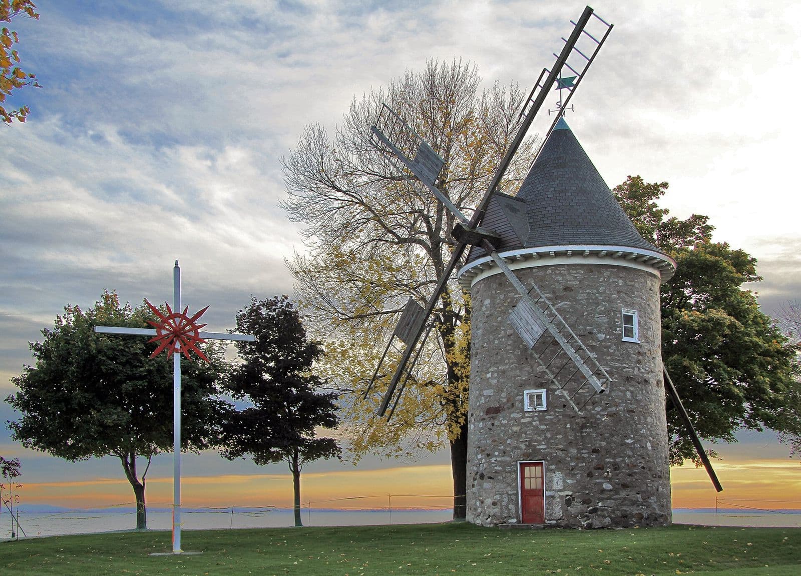 The round stone windmill of Pointe-Claire, with its red door and wooden sails, on a lawn by Lac Saint-Louis under an evening sky